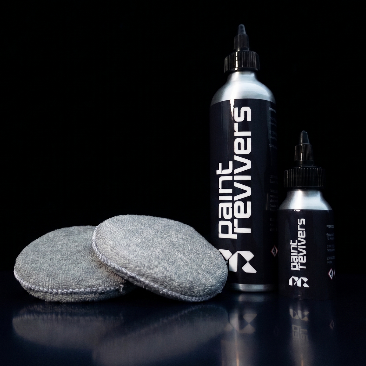 Two gray circular felt pads with stitched edges stand in front of two Paint Revivers bottles on a glossy black surface against a black background.